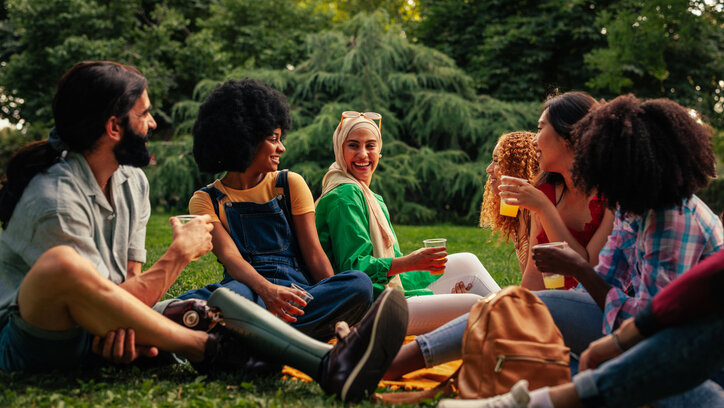A group of diverse friends are on a day time party in the park, chatting, sitting, drinking an laughing.